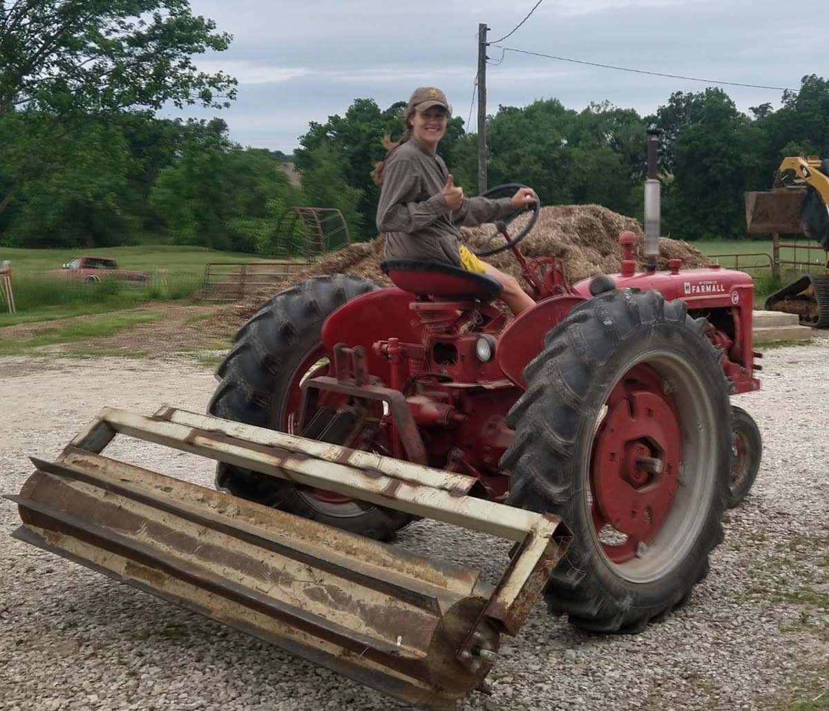 Elizabeth Leonard on a red Farmall tractor in the farmyard, giving a thumbs up